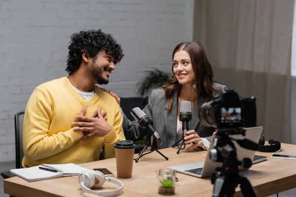 Happy indian podcaster talking to smiling colleague near microphones, notebooks, headphones, smartphone with blank screen and digital camera on blurred foreground in broadcasting studio — Stock Photo