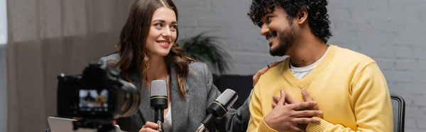Honest and bearded indian man touching chest and talking to charming brunette colleague near microphones and blurred digital camera in radio studio, banner — Stock Photo