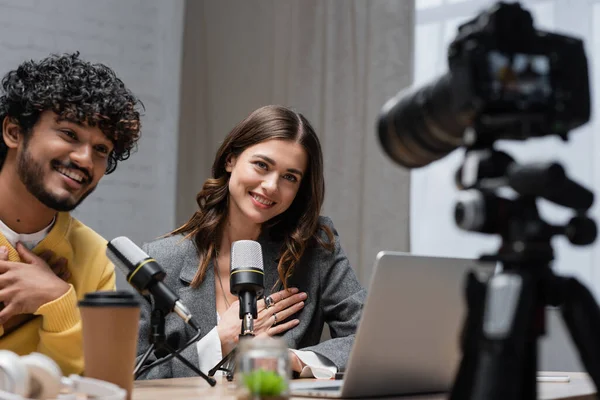 Pleased multiethnic radio hosts looking at blurred digital camera near microphones and takeaway drink while recording podcast in professional radio studio — Stock Photo