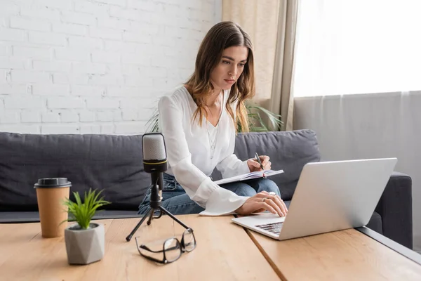 Brunette woman in white blouse using laptop and writing in notebook near microphone, coffee to go, flowerpot and eyeglasses on table in professional radio studio — Stock Photo