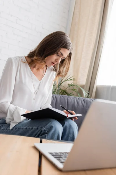 Charmante und nachdenkliche Frau in weißer Bluse, Stift in der Hand und Blick in Notizbuch, während sie auf der Couch neben verschwommenem Laptop im Sendestudio sitzt — Stockfoto