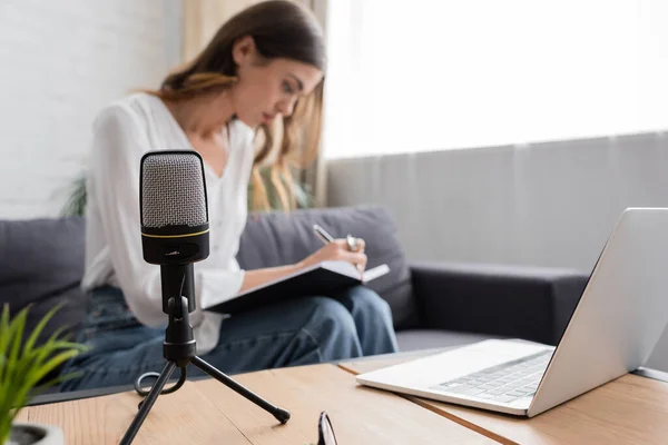 Focus on professional microphone and laptop on table near blurred charming broadcaster writing in notebook while sitting on couch in modern studio — Stock Photo