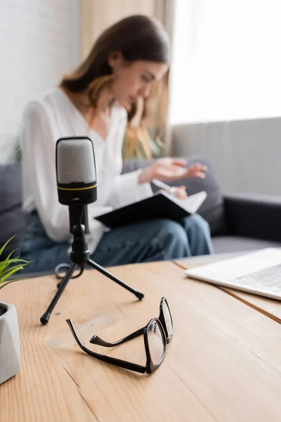 Selective focus of table with laptop and eyeglasses and professional microphone near podcaster sitting on couch and working with notebook on blurred background — Stock Photo