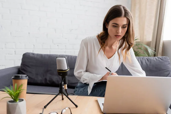 Pensive brunette woman sitting on couch and looking at laptop while preparing podcast near professional microphone, takeaway drink and flowerpot on table in radio studio — Stock Photo
