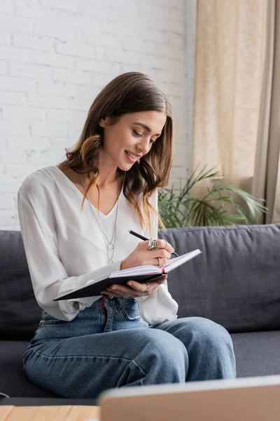 Cheerful and pretty broadcaster with brunette hair wearing white blouse and writing in notebook with pen while sitting on couch and preparing podcast in radio studio — Stock Photo