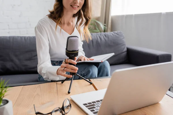 Cropped view of smiling woman in white blouse and blue denim jeans sitting on couch with notebook and adjusting microphone near eyeglasses on table in radio studio — Stock Photo