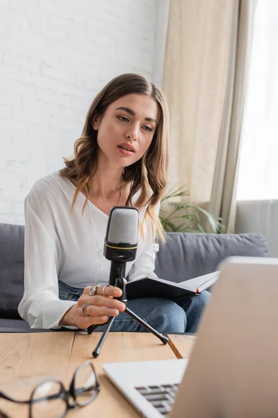 Charming and thoughtful brunette woman in white blouse holding professional microphone and notebook while sitting near laptop and blurred eyeglasses in radio studio — Stock Photo