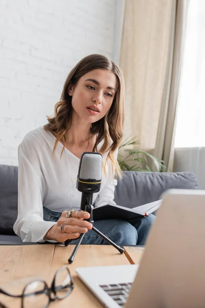 Affascinante e premurosa donna bruna in camicetta bianca con microfono e notebook professionali seduti vicino al computer portatile e occhiali sfocati in studio radio — Foto stock