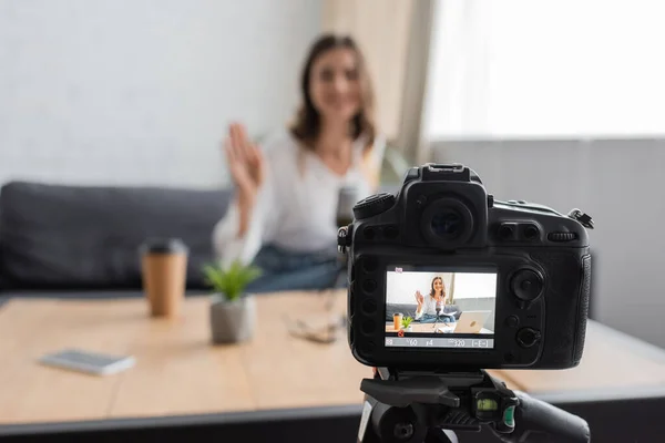 Focused photo of professional digital camera in front of blurred woman waving hand while recording podcast near paper cup, smartphone and flowerpot on table in studio — Stock Photo