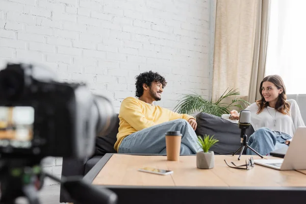 Кудрявый индиец в желтом свитере и брюнетка в белой блузке разговаривают на диване рядом с профессиональным микрофоном, ноутбуком, кофе, чтобы пойти и мобильный телефон перед размытой цифровой камерой — стоковое фото