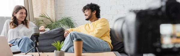 Charming brunette woman and curly indian man in yellow jumper talking on couch near microphone and blurred paper cup with flowerpot while recording podcast on blurred digital camera, banner — Stock Photo