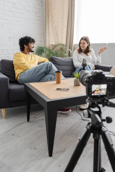 Smiling interracial couple of podcasters talking on sofa near table with microphone, paper cup, flowerpot and smartphone with blank screen in front of blurred digital camera in radio studio — Stock Photo