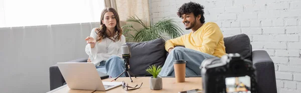 Brunette woman pointing at laptop near microphone, coffee to go and flowerpot while sitting with smiling indian podcaster in yellow jumper on sofa in radio studio, banner — Stock Photo