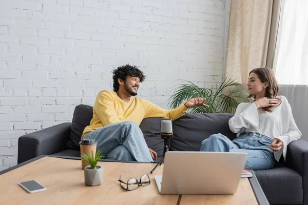 Happy indian man in yellow jumper pointing with hand while talking to colleague on sofa near laptop, flowerpot, coffee to go, eyeglasses and mobile phone with blank screen in broadcasting studio — Stock Photo