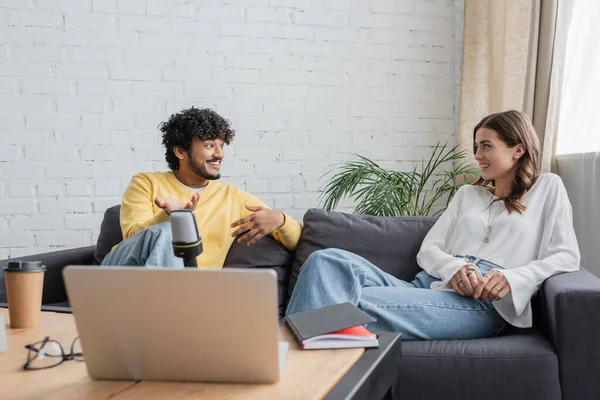 Young indian man in yellow jumper and smiling brunette woman sitting on couch near laptop, microphone, notebook, paper cup and blurred eyeglasses while talking in modern studio — Stock Photo