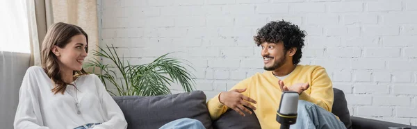 Кучерявий індійський чоловік в жовтому джемпі жестикулює, сидячи на дивані в радіостудії і розмовляючи зі усміхненою брюнеткою в білій блузці біля зеленої рослини проти цегляної стіни, банер — стокове фото