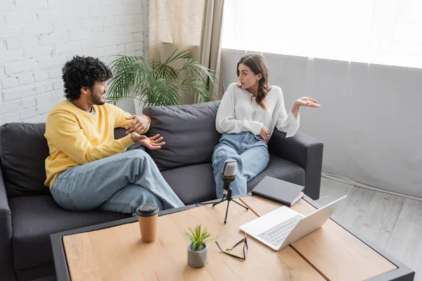 High angle view of multiethnic couple in casual clothes gesturing and talking on sofa near table with laptop, microphone, coffee to go, eyeglasses and flowerpot in radio studio — Stock Photo