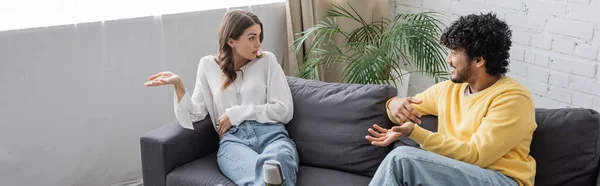 Emotional brunette woman in white blouse gesturing while talking to young and curly indian man in yellow jumper sitting on sofa near green plant against brick wall in radio studio, banner — Stock Photo
