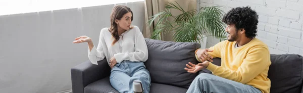 Mujer morena emocional en blusa blanca gesto mientras habla con el joven y rizado hombre indio en jersey amarillo sentado en el sofá cerca de la planta verde contra la pared de ladrillo en el estudio de radio, pancarta - foto de stock