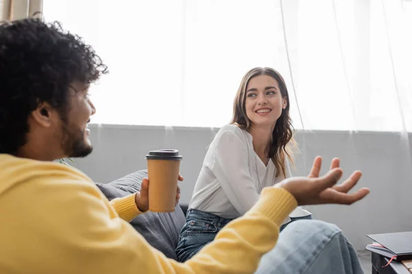 Young indian man in yellow jumper holding takeaway drink while gesturing and talking to smiling brunette woman in white blouse sitting on couch near notebook on table in radio studio — Stock Photo