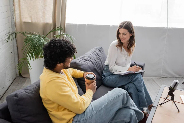 Smiling brunette woman writing in notebook while sitting on couch near curly indian man in yellow jumper holding coffee to go near professional microphone in radio studio — Stock Photo
