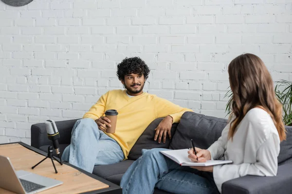 Brunette woman writing in notebook near curly and smiling indian man in yellow jumper holding coffee to go while talking on couch near microphone and laptop in radio studio — Stock Photo