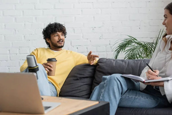 Smiling indian man in yellow jumper holding coffee to go and talking to woman writing in notebook near laptop and microphone while recording podcast in radio studio — Stock Photo