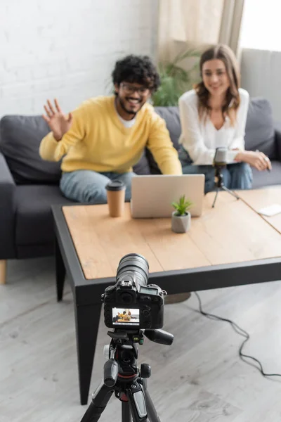 Blurred indian man waving hand during video chat on laptop near smiling colleague and professional digital camera in broadcasting radio studio with paper cup and flowerpot on table — Stock Photo