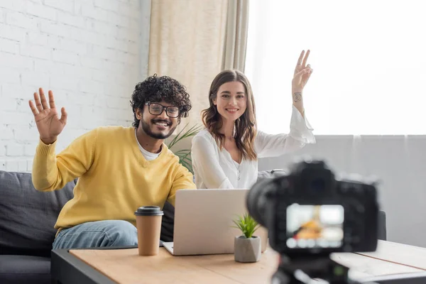 Cheerful interracial vloggers in casual clothes waving hand and showing victory gesture during video call on laptop near blurred digital camera in radio studio — Stock Photo