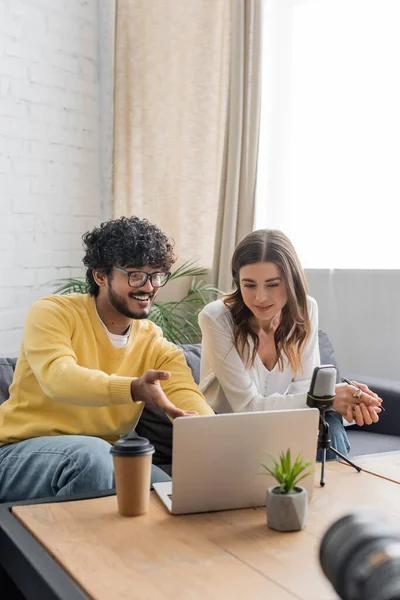 Happy indian podcaster in eyeglasses and yellow jumper pointing at laptop near brunette colleague, microphone and paper cup while having video call in front of blurred digital camera in studio — Stock Photo