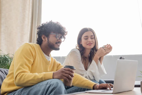 Smiling indian broadcaster in eyeglasses and yellow jumper holding coffee to go and using laptop near charming colleague during video call in radio studio — Stock Photo