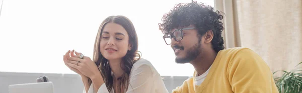 Charmante femme brune et bouclée homme indien en lunettes et pull jaune regardant ordinateur portable flou et souriant pendant l'appel vidéo en studio podcast, bannière — Photo de stock