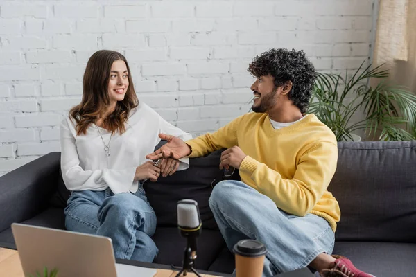 Interracial radio hosts looking at each other and talking while sitting on sofa near laptop and professional microphone with coffee to go while recording podcast in studio — Stock Photo