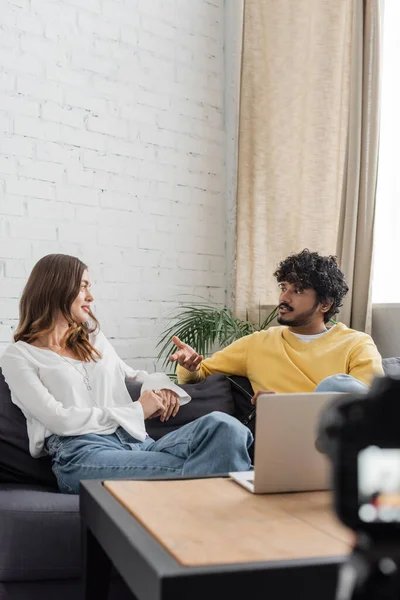 Young and curly indian man in yellow jumper gesturing and talking to charming brunette woman in white blouse white sitting on couch near laptop while recording podcast in studio — Stock Photo