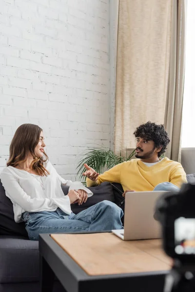 Jeune homme indien bouclé en pull jaune gesticulant et parlant à une charmante femme brune en chemisier blanc assise sur un canapé près d'un ordinateur portable tout en enregistrant un podcast en studio — Photo de stock