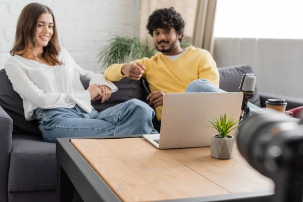 Smiling indian podcaster pointing at laptop while sitting on couch near charming and happy colleague during video chat near microphone, coffee to go and flowerpot in front of blurred digital camera in studio — Stock Photo