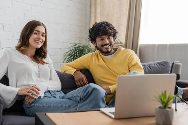 Cheerful multiethnic couple of podcasters sitting on couch and looking at laptop during video chat near blurred microphone and flowerpot with tiny plant on table in studio — Stock Photo
