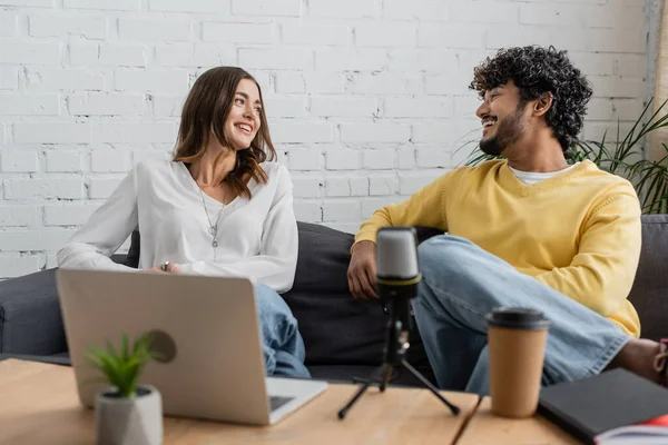 Joyful interracial podcasters smiling at each other during conversation near blurred laptop and professional microphone while sitting together on grey couch in studio — Stock Photo