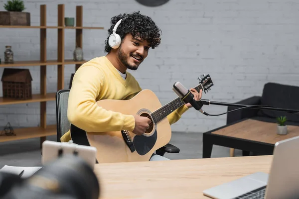 Gioioso musicista indiano in cuffia e maglione giallo che suona la chitarra acustica durante la registrazione di podcast vicino al microfono professionale e laptop sfocato con fotocamera digitale in studio — Foto stock