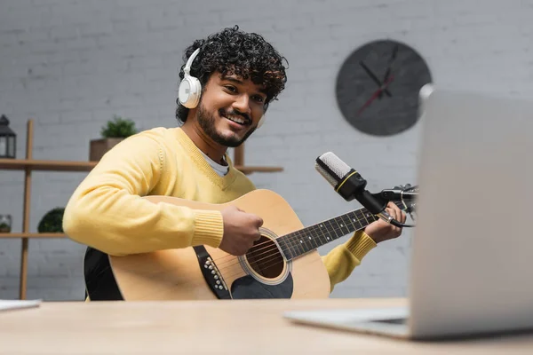 Молодой и веселый индийский музыкант в наушниках и желтый джампер играет на акустической гитаре во время записи музыки в студии рядом с профессиональным микрофоном и размытым ноутбуком — стоковое фото