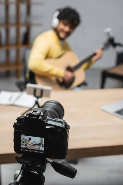Foco em podcast de gravação de câmera digital profissional perto de smartphone em tripé, músico indiano tocando guitarra acústica em fundo embaçado em estúdio — Fotografia de Stock