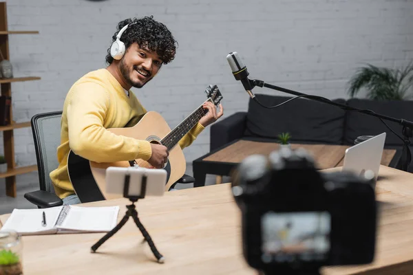 Happy indian musician in headphones and yellow jumper playing acoustic guitar near smartphone on tripod, laptop, notebook and digital camera on blurred foreground in radio studio — Stock Photo