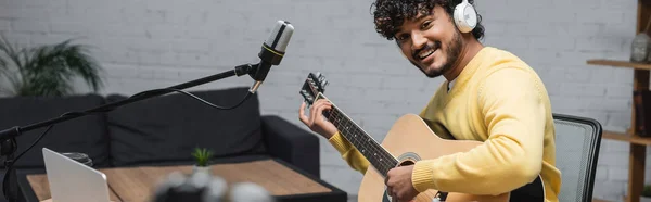 Кудрявый и счастливый индийский музыкант в наушниках и желтый джампер играет на акустической гитаре возле ноутбука и профессиональный микрофон в студии с диваном на заднем плане, баннер — стоковое фото