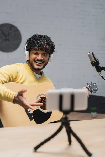 Homem indiano feliz em fones de ouvido e jumper amarelo sentado com guitarra acústica e gesto perto de smartphone borrado no tripé e microfone profissional em estúdio de gravação — Fotografia de Stock