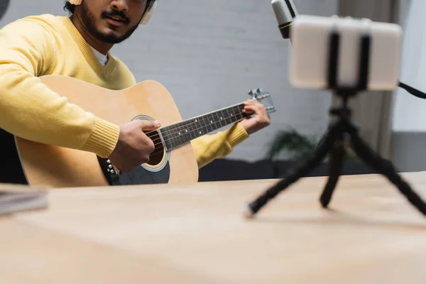 Vista recortada del músico indio creativo en puente amarillo tocando la guitarra acústica cerca del micrófono y el teléfono inteligente con trípode mientras graba podcast en primer plano borroso - foto de stock