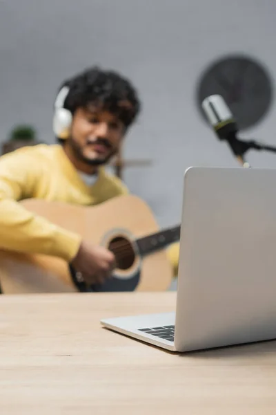 Фокус на ноутбуці на дерев'яному столі біля молодого індійського чоловіка в бездротових навушниках, який грає на акустичній гітарі під час запису подкасту біля мікрофона в радіостудії — стокове фото