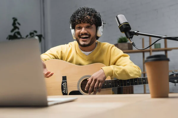 Alegre músico indio en auriculares y puente amarillo tocando la guitarra acústica cerca borrosa portátil y micrófono de estudio en la mesa con café borroso para llevar mientras se graba podcast en el estudio - foto de stock