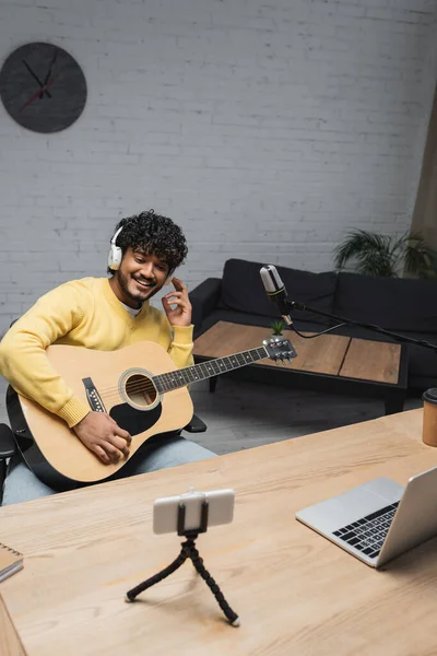 Улыбающийся молодой индийский вещатель в беспроводных наушниках, держащий акустическую гитару рядом с микрофоном, смартфоном и ноутбуком во время потока в студии — стоковое фото