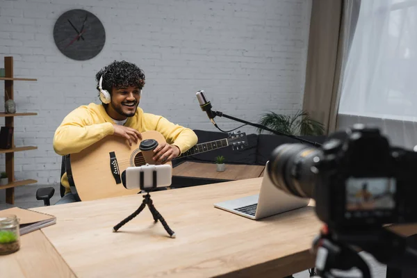 Sonriente podcaster indio en auriculares que sostienen la guitarra acústica y el café para acercarse al micrófono y los dispositivos durante la transmisión en el estudio de podcast - foto de stock