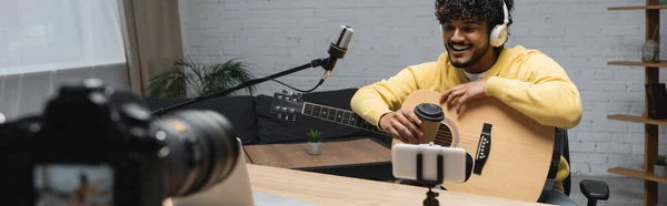 Веселый молодой индийский блогер в беспроводных наушниках держит кофе, чтобы подойти к акустической гитаре, микрофону и устройствам в студии, баннер — стоковое фото